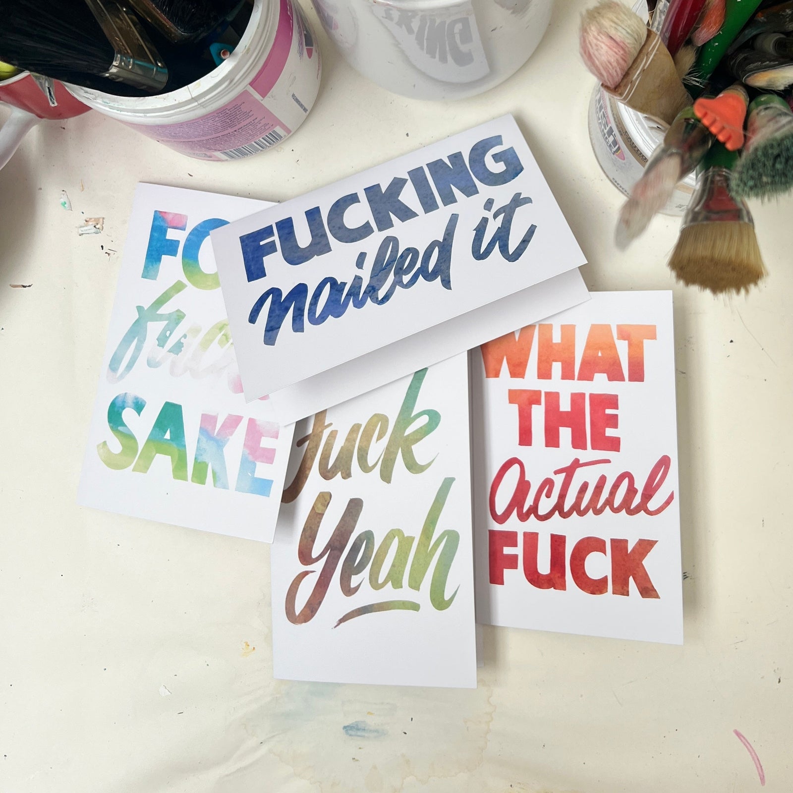 Art supplies including paintbrushes and colorful cards with profane text on a white surface.